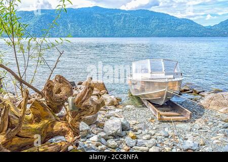 Blick auf die Wurzel eines alten Haken ein Sinken Baum und ein Motorboot auf dem Hintergrund eines see mit einer Berglandschaft Stockfoto