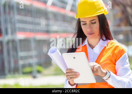 Junge Frau in PSA hält eine Tablette und eine Blaupause mit einer Baustelle im Hintergrund. Stockfoto