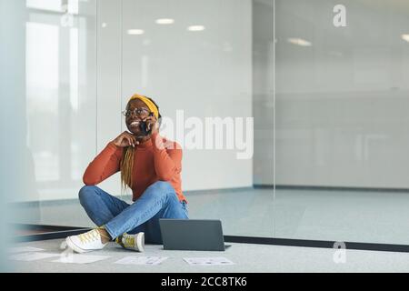 Afrikanische Designerin sitzt auf dem Boden mit Laptop und Telefonieren auf dem Handy in leerem Büro Stockfoto