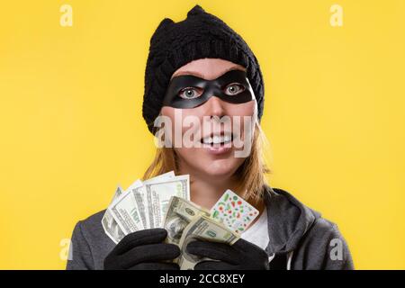 Cyberkriminalität und Gelddiebstahl. Eine Frau mit Hut, Handschuhen und Maske hält einen Geldwappen in den Händen und schaut träumerisch auf. Gelber Hintergrund. Speicherplatz kopieren. Stockfoto