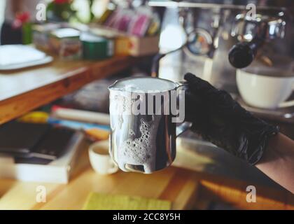 Barista trägt medizinische Latexhandschuhe, bereitet Kaffeegetränk mit Espressomaschine im Shop Café zu. Frau hält den Krug mit Metallmilch in der Hand. Stockfoto