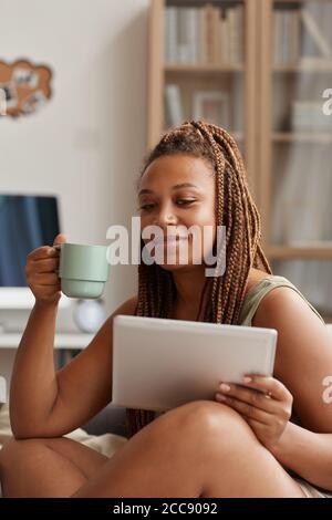 Afrikanische junge Frau sitzt auf dem Sofa mit einer Tasse Kaffee Und mit einem digitalen Tablet zu Hause Stockfoto