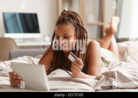 Junge Frau auf dem Bett liegend mit digitalen Tablet und Kredit Karte Sie kauft Kleidung im Online-Shop Stockfoto
