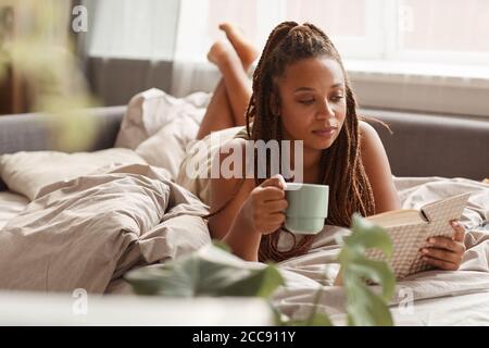 Junge Frau liegt im Bett trinken Kaffee und lesen ein Buchen Sie am Morgen, sie genießt ihre Wochenenden Stockfoto