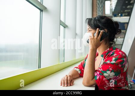 Frau am Flughafen trägt chirurgische Schutzmaske, Gespräch am Telefon lächelnd und positiv, Reisen während der covid-19 Pandemie. Das Virus hat ca. Stockfoto
