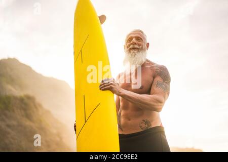 Tätowierte Senioren-Surfer, die bei Sonnenuntergang Surfbrett am Strand halten - fröhlicher alter Kerl, der Spaß hat, Extremsport zu machen - freudiges älteres Konzept - Fokus auf Hi Stockfoto