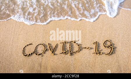 Covid-19 geschrieben auf dem Sand des Küstenstrandes mit eingehender Wellenwaschung, Löschen oder Löschen es während Coronavirus Sommer. Angst vor dem Urlaub wegen Stockfoto