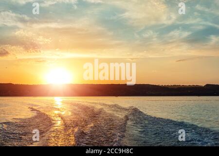 Szenische helle lebendige blau bis rot warmen Sonnenuntergang Abend Landschaft mit Motorboot Wirbel Spur auf Wasseroberfläche des Sees oder des Flusses. Horizont-Küstenlinie Stockfoto