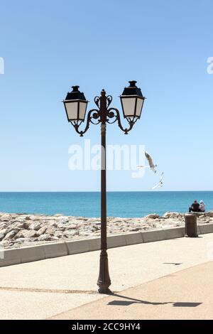 Laterne auf einer Promenade am mittelmeer, zwei nicht identifizierbare Personen sitzen auf den Felsen, zwei Möwen fliegen Stockfoto