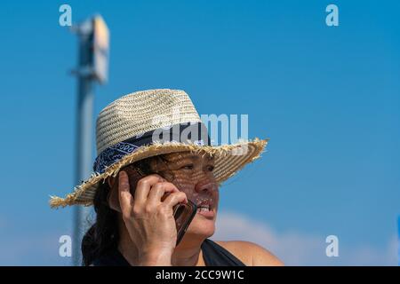 Nahaufnahme Bild einer asiatischen Frau mittleren Alters mit einem Strohhut sprechen in einem Smartphone einen heißen Sommertag. Strahlend blauer Himmel im Hintergrund Stockfoto