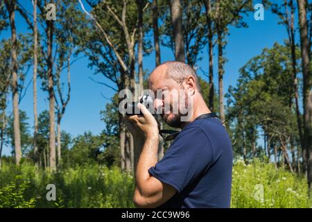 Erwachsene Mann hält spiegellose Kamera, Aufnahmen in einem Wald im Sommer Tag, Halbkörperporträt Stockfoto