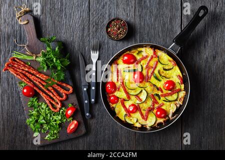 Omelette mit Zucchini, dünnen Räucherwürsten und Tomaten in einer Pfanne auf einem dunklen Holztisch mit Zutaten auf einem Schneidebrett, horizontale Ansicht Fr. Stockfoto
