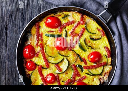 Frittata, Omelette mit Zucchini, dünne Räucherwürste und Tomaten, die in einer Pfanne auf einem dunklen Holztisch gefüllt sind, horizontale Ansicht von oben, flach liegend, c Stockfoto