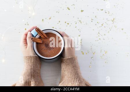 Heiße Schokolade mit Zimt Gewürz in Emaille Becher in weiblichen Händen auf weißem Holz Hintergrund Draufsicht. Heißer gemütlicher Drink für Herbst oder Winter. Stockfoto