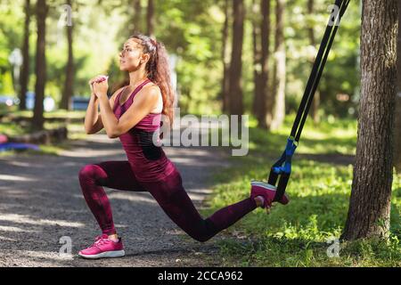 Junge Frau macht ein Bein hocken mit Fitness-Gurte an einem Baum im Park befestigt. Gesundes Lifestyle-Konzept Stockfoto