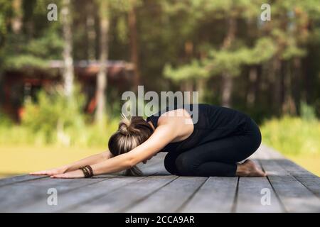 Frau in Sportkleidung sitzt auf einer Holzbrücke praktiziert Yoga Asanas, Durchführung einer Kinderpose Stockfoto