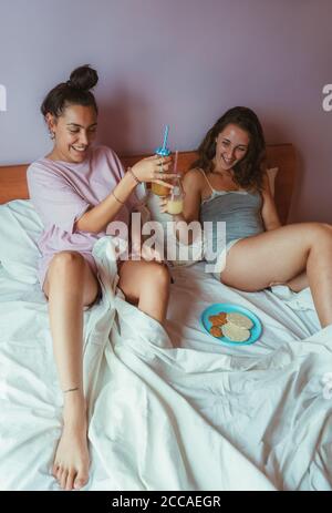 Zwei Freundinnen frühstücken im Bett des Hotelzimmers, liegen zusammen und unterhalten sich. Stockfoto