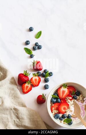Sommer Frühstück. Schüssel Joghurt mit Erdbeeren, Heidelbeeren und Müsli auf weißem Marmorhintergrund mit beigem Leinen und mehr Obst. Stockfoto
