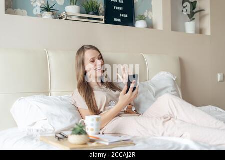 Glücklich und lächelnd entspannte fröhliche Frau mit Smartphone und machen Selfie auf dem Bett mit Kaffee. Blonde Mädchen mit Handy und mit einem Video Stockfoto