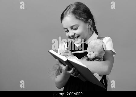 Mädchen hält Teddybären und schreibt in Blau notebook. Zurück zu Schule und Bildung Konzept. Schule Mädchen mit glücklichen Gesicht auf blauem Hintergrund isoliert. Schüler in der Schule Uniform mit Zopf und rosa Rucksack Stockfoto