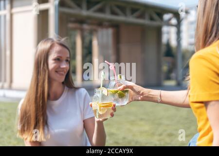 Zwei schöne junge Mädchen in der sonnigen Stadt im Sommer trinken einen Cocktail aus Gläsern. Sitzen auf Gras im Park. Zwei lachende Freunde entspannen sich mit lemo Stockfoto