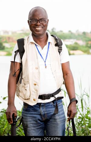 Reifer Mann, der am Rand des Wassers in beige gekleidet steht, Brille, Rucksack lächelnd auf die Kamera. Stockfoto