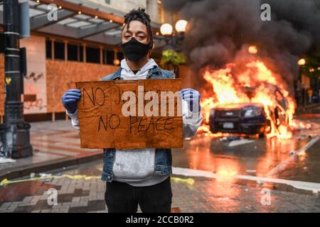 Seattle, USA, 30. Mai 2020: Ein Aktivist hält ein Zeichen NO JUSTICE NO PEACE, während ein Polizeiauto in Westlake im Hintergrund brennt. Stockfoto