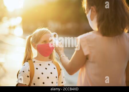 Moderne Lehrerin und Schülerin mit Maske misst die Temperatur mit einem Digitalthermometer im Freien. Stockfoto