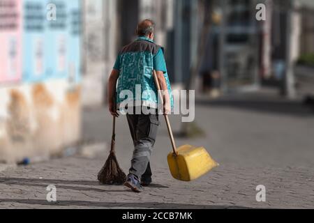 Ein Reinigungsarbeiter mit Besen und Kehrschaufel, der auf einer abgeschiedenen Straße läuft, isoliert und mit verschwommenem Hintergrund hervorgehoben. Stockfoto