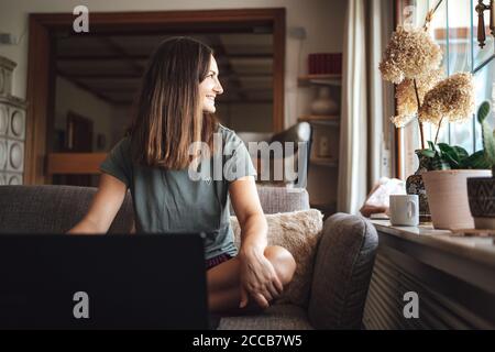 Lächelnde junge Frau mit braunen Haaren sitzt mit Laptop auf Sofa zu Hause Stockfoto