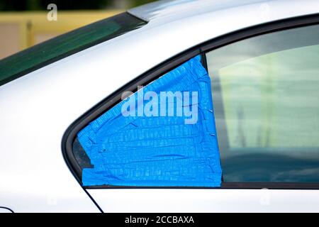 Kaputtes und beschädigtes Autofenster, das vorübergehend mit blauem Klebeband bedeckt ist, um das Innere vor Regen und Wasser zu schützen. Smash-and-Grab Automobil-Einbrüche. Stockfoto