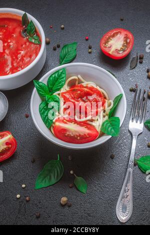 Spaghetti mit Sauce auf einem Teller und Tomatensauce mit Basilikum in einer kleinen Schüssel mit Gewürzwürzen auf einem schwarzen Tisch. Kopierraum, vertikale Ausrichtung, Stockfoto