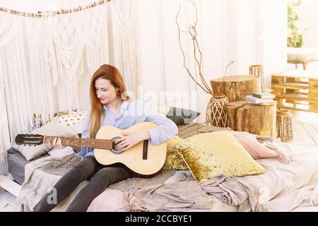 Eine Erwachsene Frau spielt die Gitarre auf dem Bett in einem Boho-Stil Zimmer sitzen. Schöne und gemütliche Atmosphäre. Stockfoto