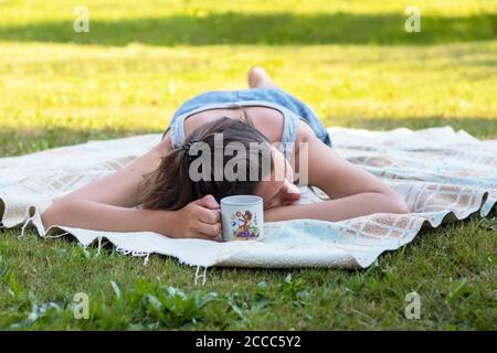 Junge schöne Frau, die auf dem Bauch auf einer Decke liegt und eine Tasse in der Hand hält, auf dem Gras, sonniger Sommertag Stockfoto
