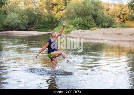 Glückliche ältere Frau in einem flachen Fluss, der mit Wasser spielt. Reife Frau enyoing in einem Fluss, spritzt Wasser im Sommer sonnigen Tag Stockfoto