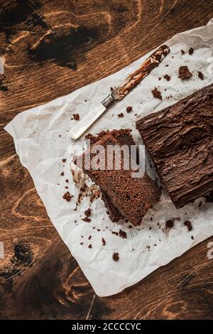 In Scheiben geschnittener Schokoladenkuchen und ein Messer auf Backpapier Auf dunklem Holzhintergrund Stockfoto