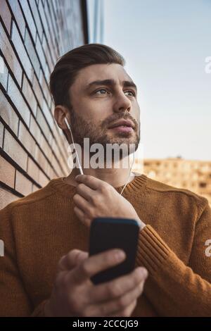 Lächelnder Mann, der Musik auf Kopfhörern hört und sich an die Ziegelwand lehnt. Stockfoto