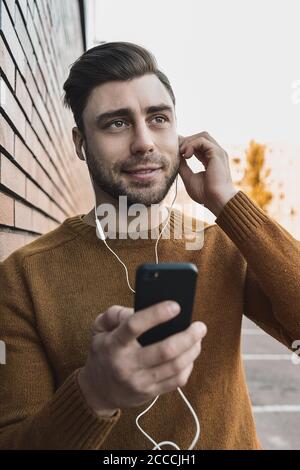 Lächelnder Mann, der Musik auf Kopfhörern hört und sich an die Ziegelwand lehnt. Stockfoto