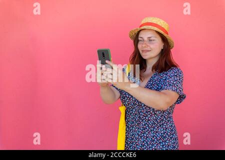 Schöne junge rothaarige Mädchen in einem Hut und Sommerkleid Steht in der Nähe einer rosa Wand in der Straße nehmen ein Selfie Stockfoto