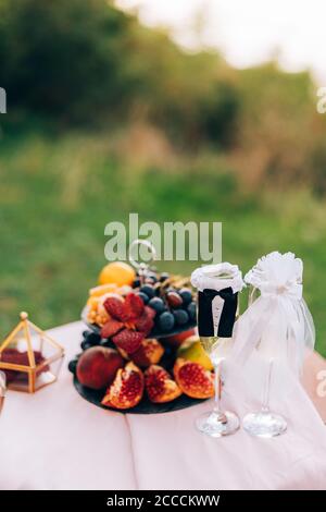 Ein bemalter Tisch mit einem Bücherregal mit Früchten und Gläsern für Champagner in den Anzügen der Braut und des Bräutigams. Stockfoto