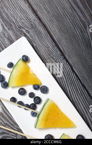 Stücke von gelber Wassermelone auf Stöcken. Auf einen Teller legen. In der Nähe sind Heidelbeeren. Platte auf bemalten Kiefernbrettern. Stockfoto