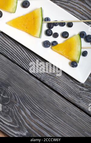 Stücke von gelber Wassermelone auf Stöcken. Auf einen Teller legen. In der Nähe sind Heidelbeeren. Platte auf bemalten Kiefernbrettern. Stockfoto