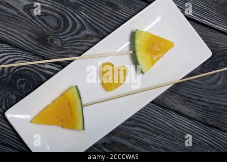 Stücke von gelber Wassermelone auf Spieße. Auf einen Teller legen. Platte auf bemalten Kiefernbrettern. Stockfoto