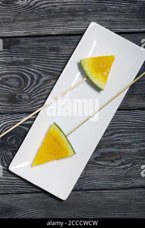 Stücke von gelber Wassermelone auf Spieße. Auf einen Teller legen. Platte auf bemalten Kiefernbrettern. Stockfoto