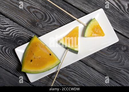 Stücke von gelber Wassermelone auf Spieße. Auf einen Teller legen. Platte auf bemalten Kiefernbrettern. Stockfoto