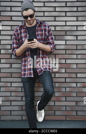 Hipster sms SMS SMS-SMS-Handy-App in der Stadt Straße auf Backsteinmauer Hintergrund. Erstaunlich Mann hält Smartphone in smart Casual wear stehen. Stockfoto