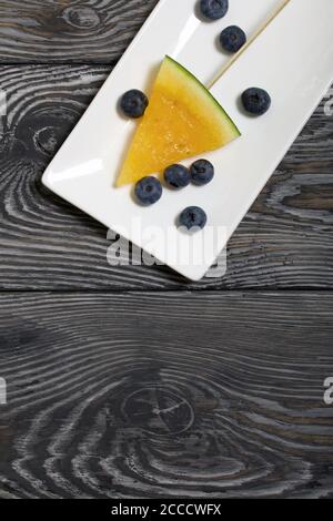 Stücke von gelber Wassermelone auf Stöcken. Auf einen Teller legen. In der Nähe sind Heidelbeeren. Platte auf bemalten Kiefernbrettern. Stockfoto