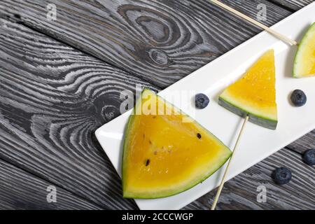 Stücke von gelber Wassermelone auf Stöcken. Auf einen Teller legen. In der Nähe sind Heidelbeeren. Platte auf bemalten Kiefernbrettern. Stockfoto