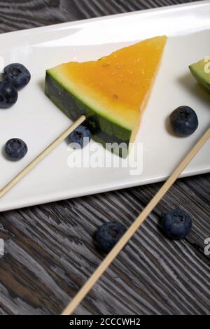 Stücke von gelber Wassermelone auf Stöcken. Auf einen Teller legen. In der Nähe sind Heidelbeeren. Platte auf bemalten Kiefernbrettern. Stockfoto