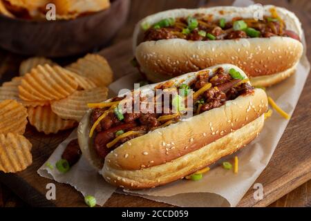 Chili Hot Dog mit Cheddar-Käse und grünen Zwiebeln auf Ein Sesambrötchen mit Kartoffelchips auf einem rustikalen Holzbrett Stockfoto